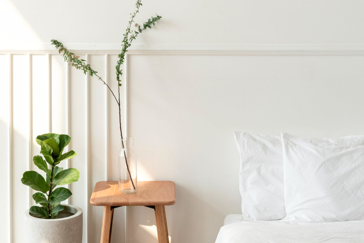 Minimalist bedroom with white bedding, a wooden side table holding a clear glass vase with a single branch, and a potted fiddle-leaf fig plant.