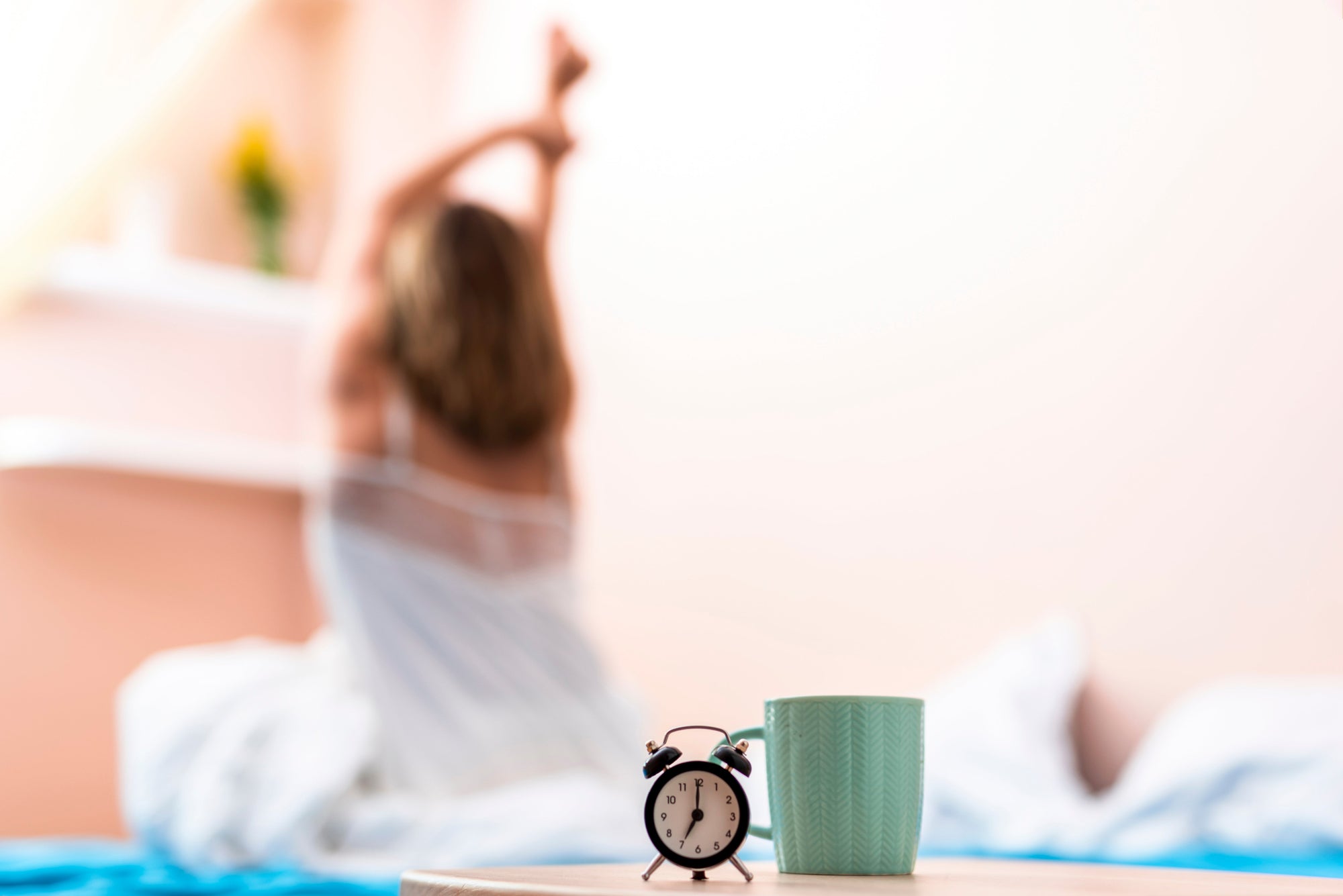 Alarm clock and coffee cup on a bedside table with a woman stretching in bed, starting her day with a reset routine in soft morning light.