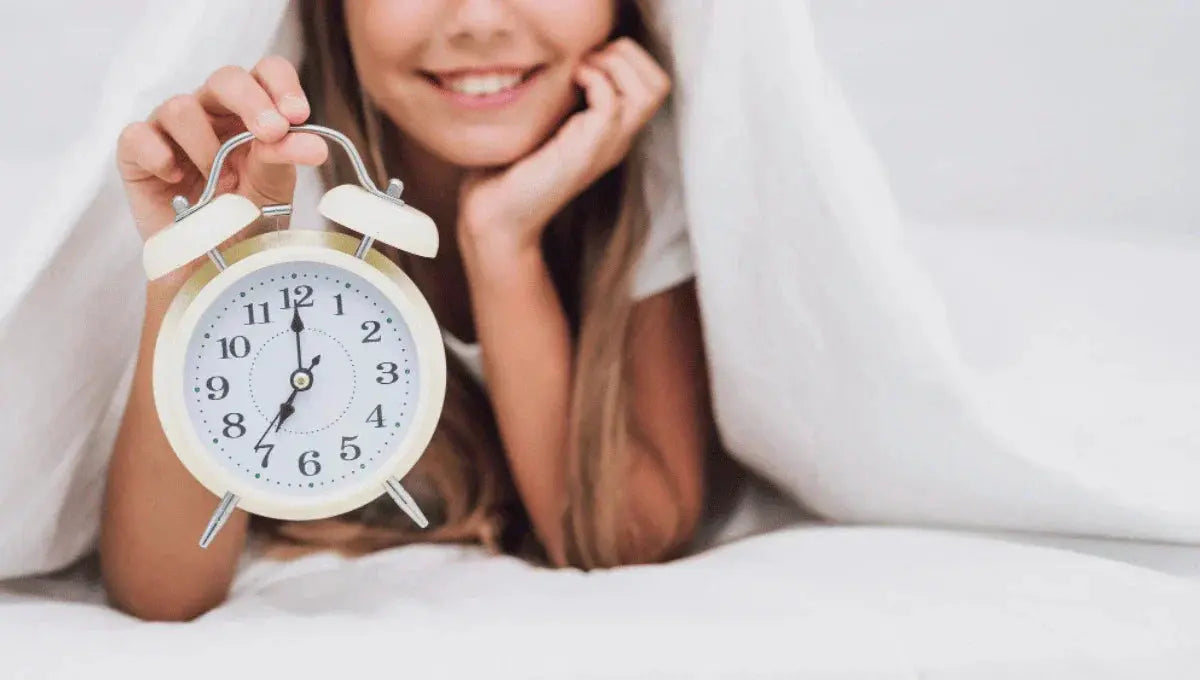 Woman in bed holding an analog alarm clock, representing adjusting sleep after daylight savings time.