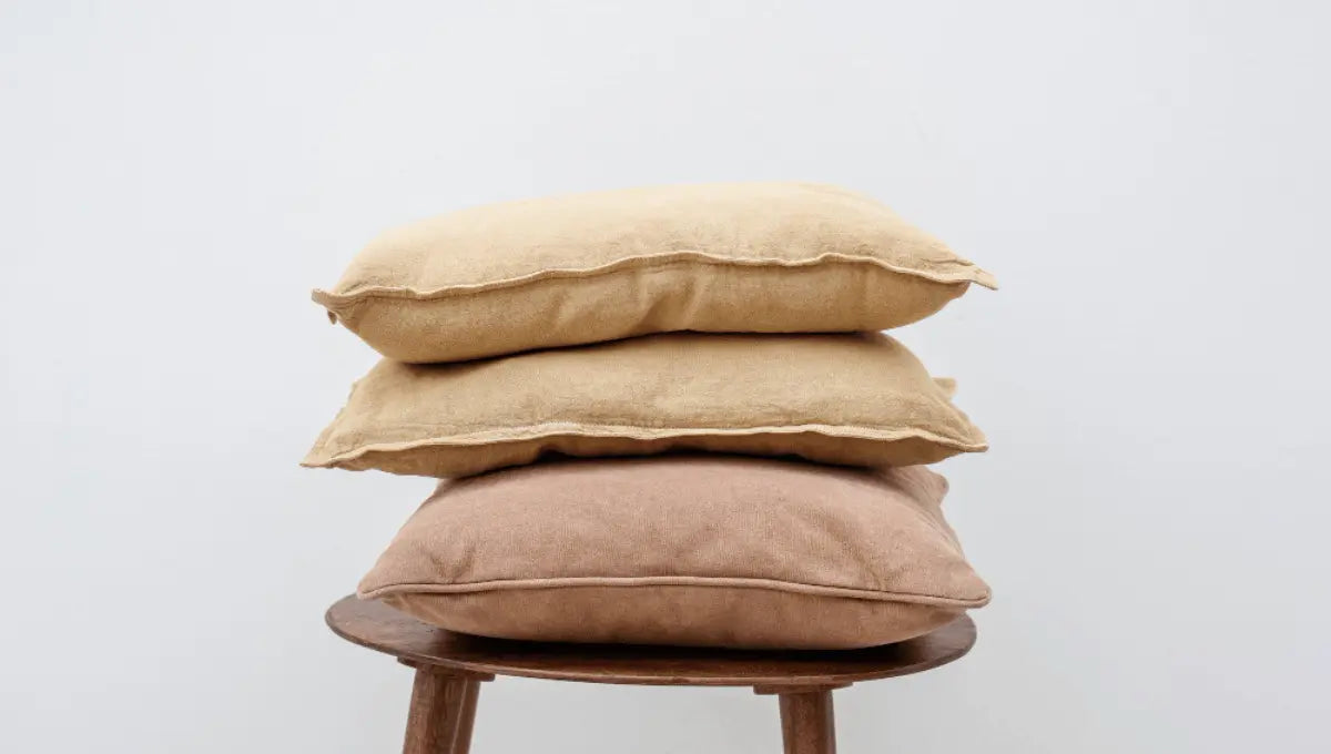 Three neutral-toned pillows stacked on a wooden stool against a soft gray wall, showing everyday pillow wear and texture.