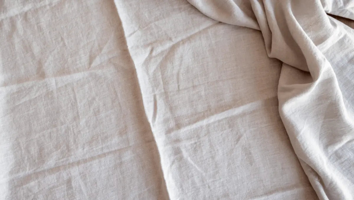 Close-up texture of light beige linen bed sheets showing natural fabric weave and soft folds.