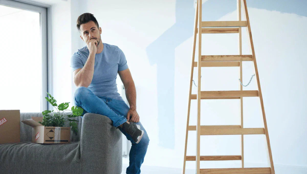 Man sitting on a couch in a partially painted living room, planning home upgrades before selling.
