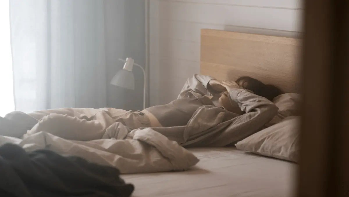 Person lying awake in bed with hand over face in softly lit bedroom during early morning light.