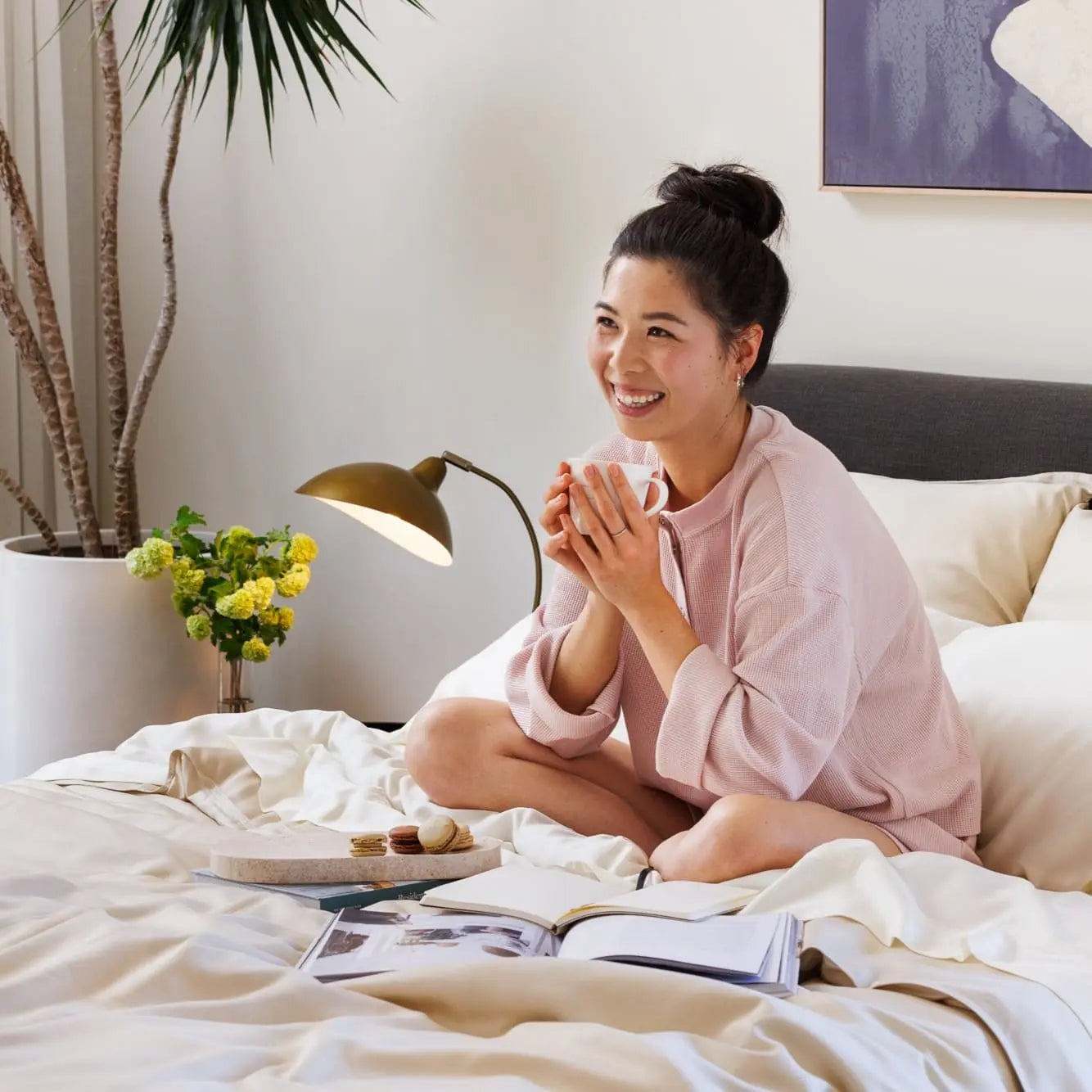 Woman sitting on a bed holding a mug, surrounded by books and a plant in a cozy room.