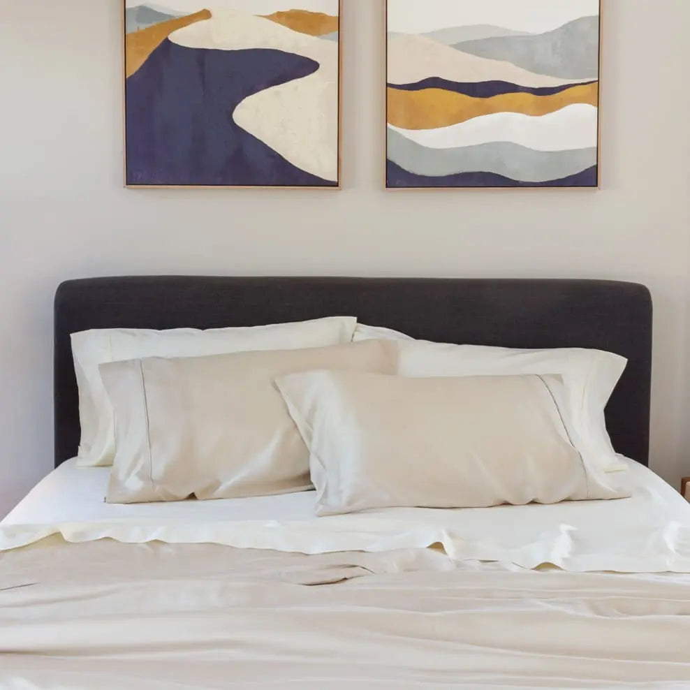 A neatly made bed with white and beige bedding, set against a dark gray upholstered headboard. Two abstract landscape wall art prints hang on the wall above the bed.