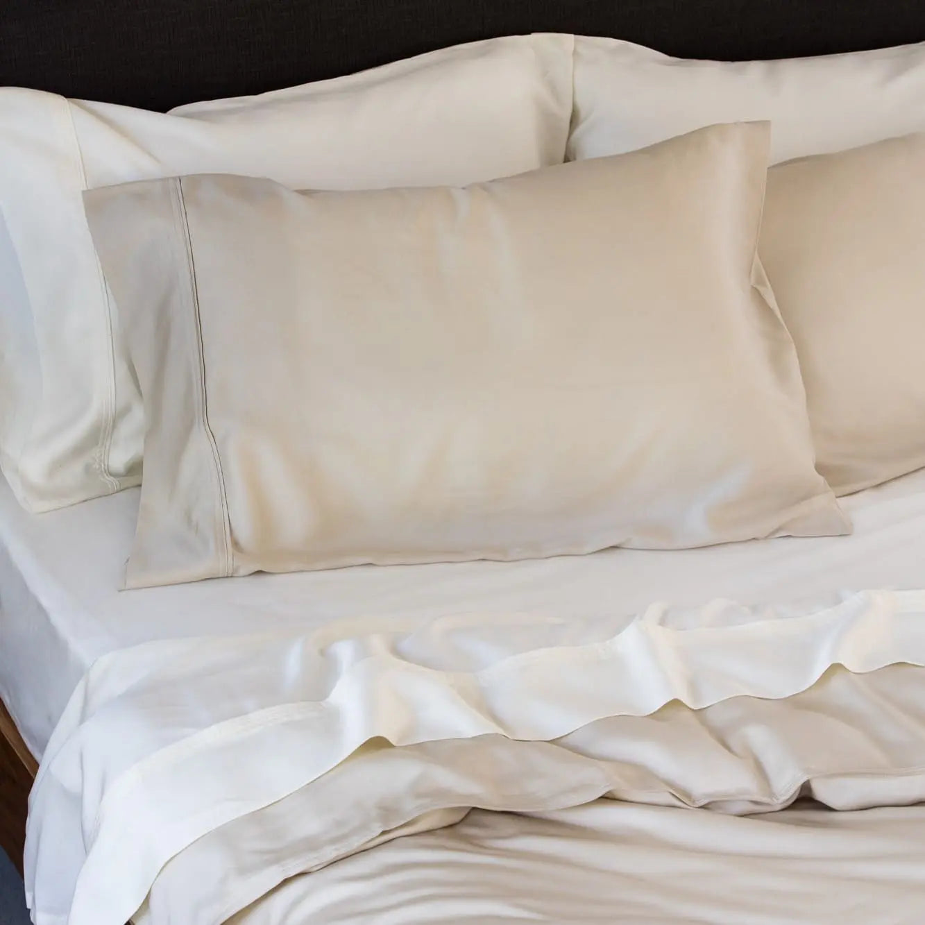 A close-up of a neatly made bed with cream-colored satin or silk pillowcases and matching white bedding, including a ruffled duvet cover.