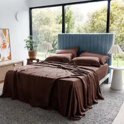 A bed dressed in dark brown bedding and matching pillows, positioned in a modern bedroom with a blue upholstered headboard, large windows showing green trees outside, and decorative side tables and plants.