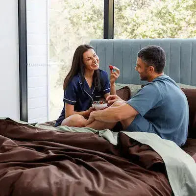 A man and a woman relax in bed on a large bed with brown satin bedding, talking and smiling in a bright bedroom with a window and a blue headboard.