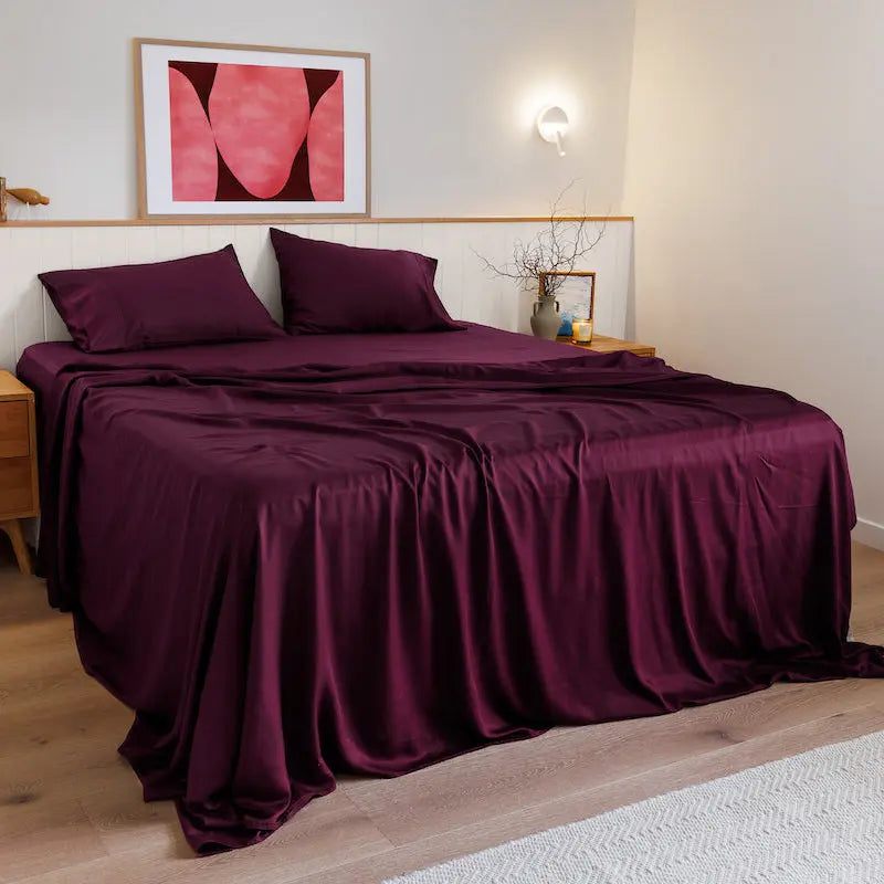 A bed dressed in rich plum purple satin bedding, including a duvet cover and two matching pillowcases, styled in a modern bedroom with light wood flooring, a wooden nightstand, and a pink abstract art print on the wall.