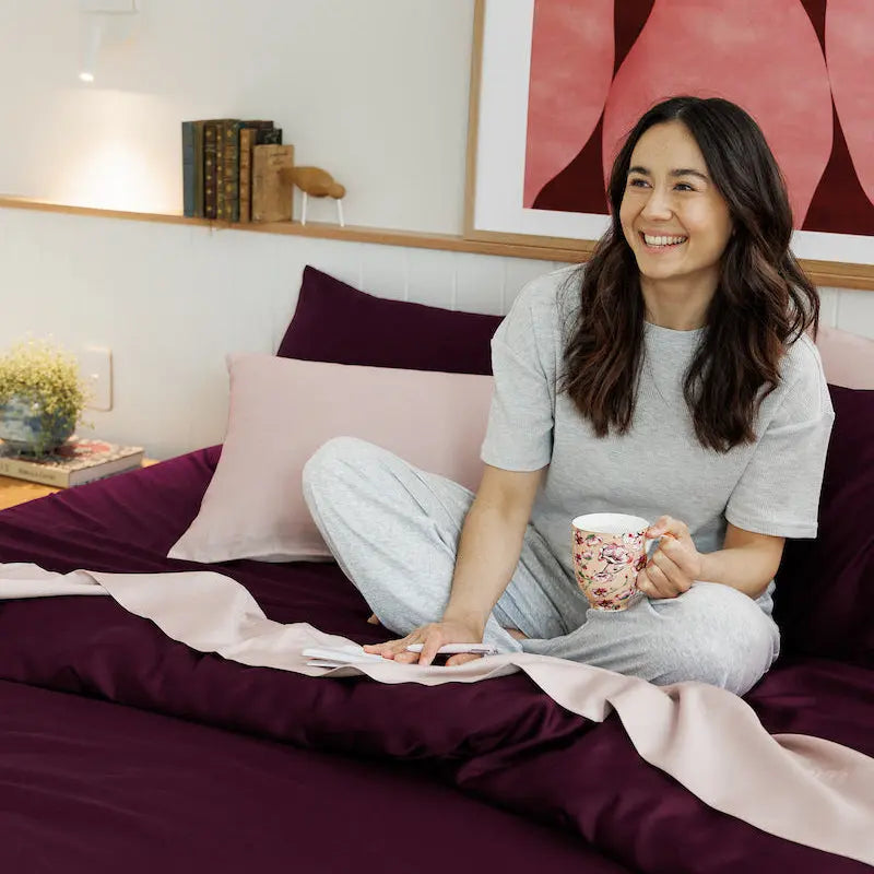 A woman sits cross-legged on a bed with deep purple bedding and a light pink blanket, holding a floral mug. She is wearing a light gray short-sleeve top and matching gray sweatpants.