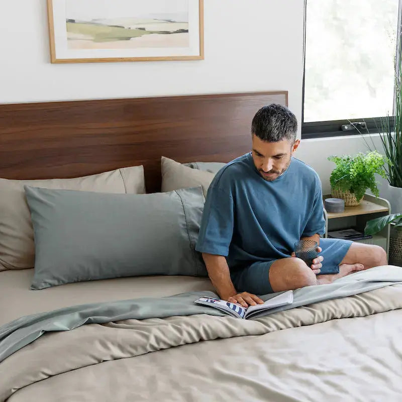 A man in a blue t-shirt and shorts sits on a bed with gray and beige bedding, reading a book in a bright, modern bedroom with plants and a wooden headboard.