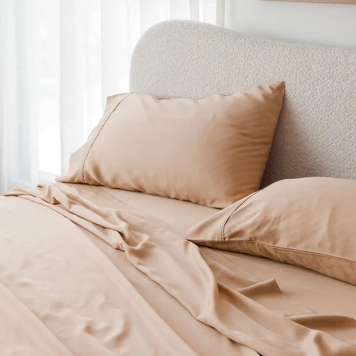 A neatly made bed with light beige or blush-beige bedding, including a duvet cover and two matching pillowcases, set against a textured cream headboard in a softly lit bedroom with sheer white curtains. #color_terracotta clay