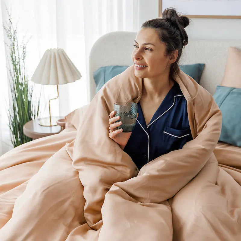 A woman in navy blue pajamas sits in bed wrapped in a large peach-colored duvet, holding a glass mug. The bedroom has white curtains, a bedside lamp, and a floral wall art print.