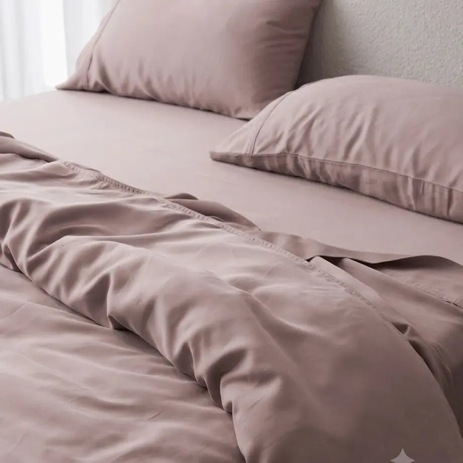 A close-up of a bed made with matching dusty mauve bedding, including a duvet cover and two pillows, styled in a softly lit bedroom.
