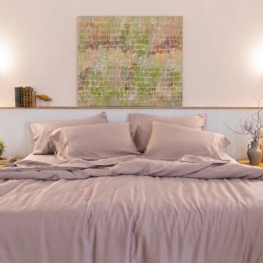 A neatly made bed with dusty mauve-lilac bedding and matching pillows, set in a bright bedroom with a light wood headboard, two bedside tables, wall sconces, and a colorful abstract painting above.
