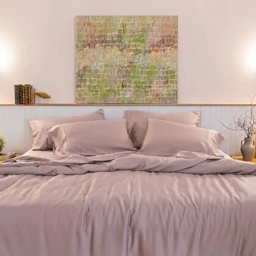 A neatly made bed with dusty mauve-lilac bedding and matching pillows, set in a bright bedroom with a light wood headboard, two bedside tables, wall sconces, and a colorful abstract painting above.