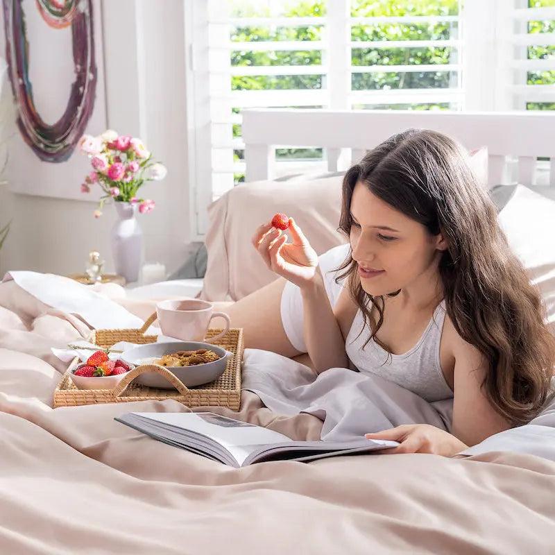 A woman with long brown hair lies on a bed reading a book, holding a strawberry. A breakfast tray with a cup, cereal, and more strawberries is placed on the bed beside her.