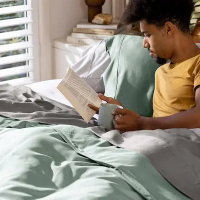 A man relaxes in bed reading a book and holding a mug, surrounded by sage green bedding in a bright, modern bedroom.