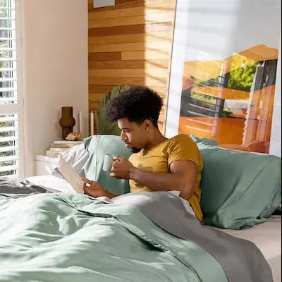 Person reading a book on a bed with green bedding in a bright room.
