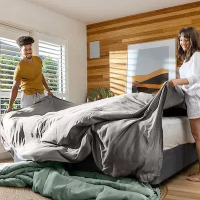 Two people setting up a bed in a bright bedroom with wooden walls and large windows.