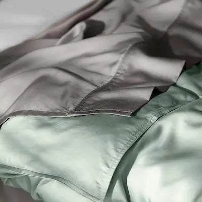 A close-up of soft, draped bedding in muted sage green and taupe-gray tones, with a visible fold and seam suggesting a duvet cover or comforter.