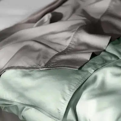 A close-up of soft, draped bedding in muted sage green and taupe-gray tones, with a visible fold and seam suggesting a duvet cover or comforter.