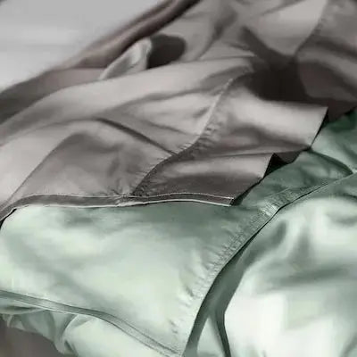A close-up of soft, draped bedding in muted sage green and taupe-gray tones, with a visible fold and seam suggesting a duvet cover or comforter.