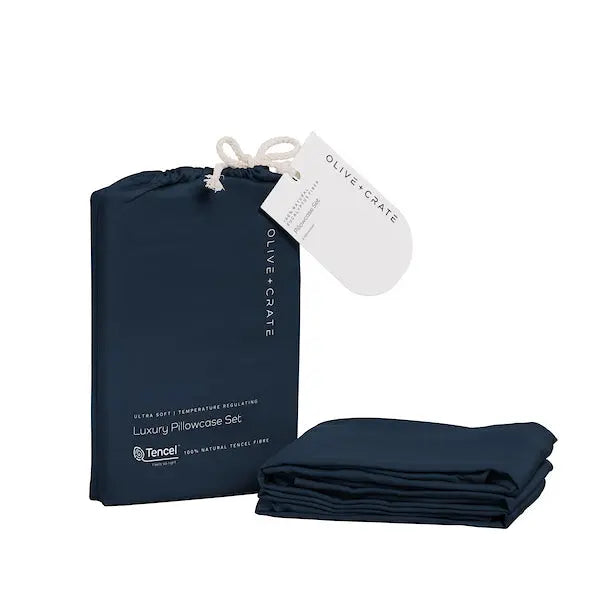 A navy blue pillowcase set shown with its branded drawstring storage bag and a folded matching pillowcase on a clean white background. The bag has a white hanging tag reading OLIVE + CRATE.