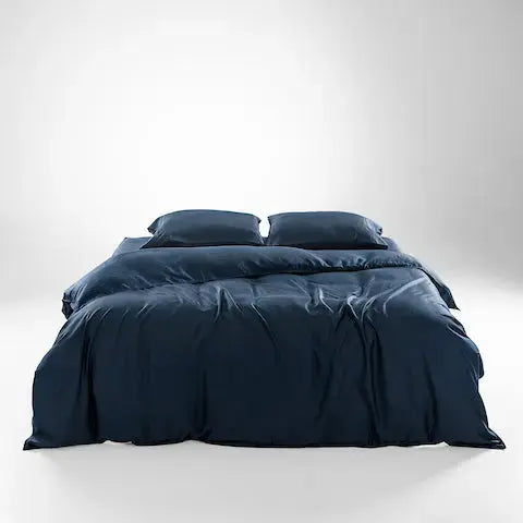A neatly made bed with dark navy blue bedding, including a duvet cover and two matching pillows, photographed in a clean studio setting with a light gray gradient background.