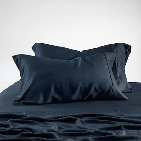 Dark navy blue bedding set with two matching pillowcases and rumpled sheets on a bed against a light gray studio background.