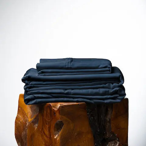 A stack of folded dark navy blue fabric, likely bedding or textiles, displayed on a rustic wooden block against a clean white studio background. French Navy sheet set