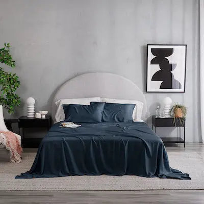A modern bedroom scene featuring a bed dressed in dark navy blue bedding, including sheets and matching pillows, set against a textured gray wall with minimalist bedside tables, a chair, and a window with sheer curtains. French Navy sheet set