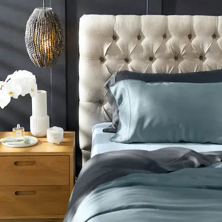 A styled bedroom scene featuring a bed with a tufted beige headboard, sage green satin bedding, and a wooden nightstand with a hanging woven pendant light against a dark gray wall.