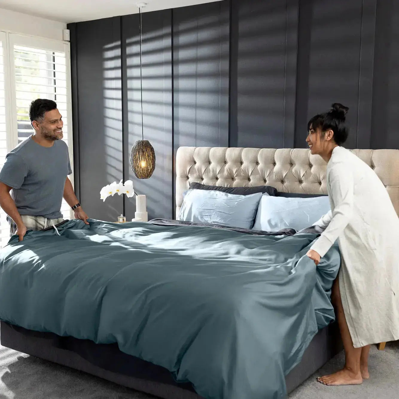 A man and a woman in a modern bedroom setting, adjusting a slate blue duvet cover on a bed with a tufted headboard. The room features dark paneled walls, hanging pendant lights, a wooden nightstand, and a window with blinds.