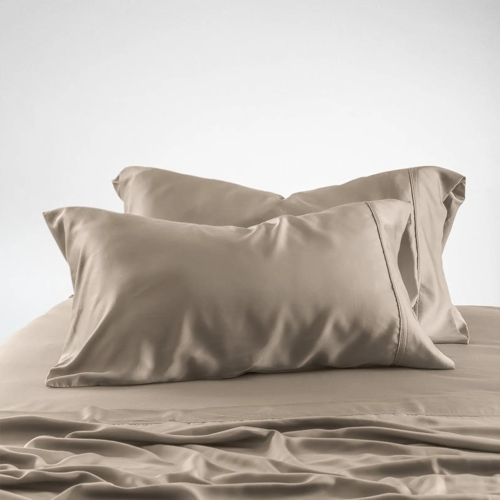 A close-up of a bed dressed in matching beige or taupe bedding, with two pillows and a wrinkled sheet against a plain light gray studio background.