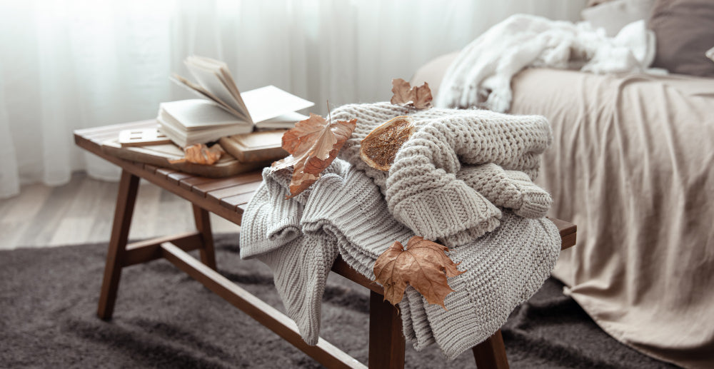 A cozy bedroom scene with folded knit sweaters and autumn leaves on a wooden bench beside an open book, creating a warm fall atmosphere for a refreshed and restful space.