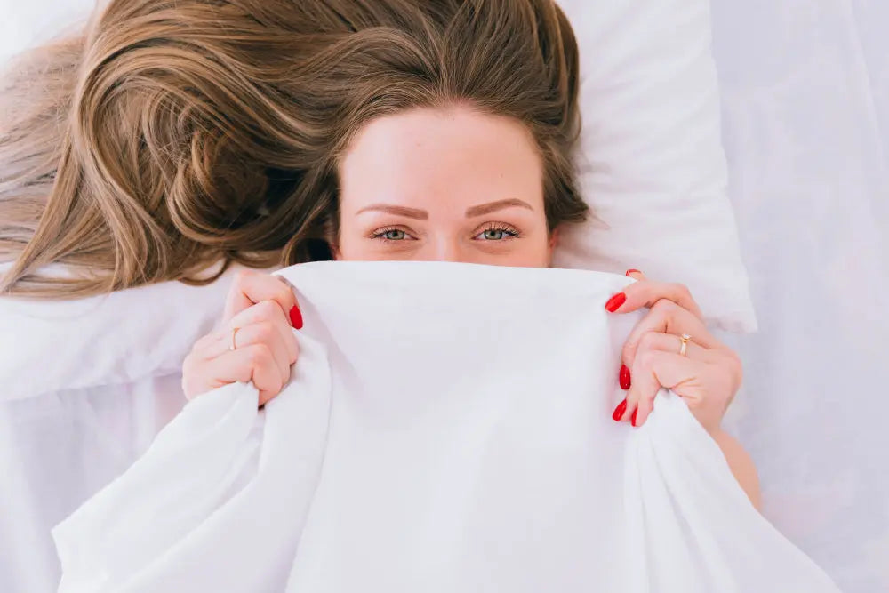 Woman with long hair smiling under white bedding, holding antimicrobial sheets up to her face.
