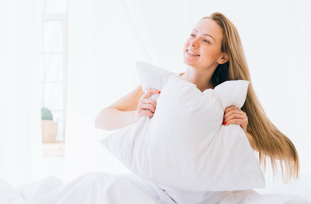 A woman sits on a bright, cozy bed smiling and hugging a soft white pillow in a peaceful, sunlit bedroom.
