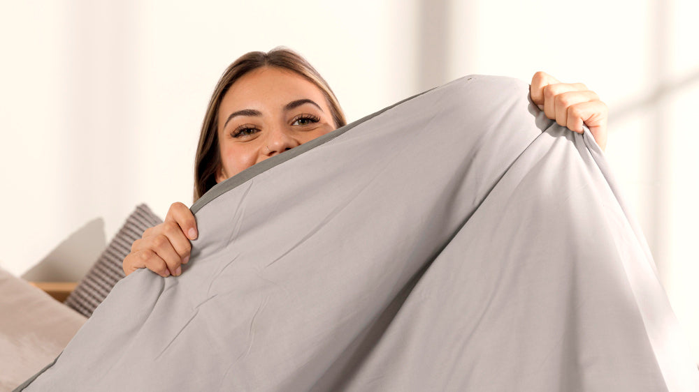 Smiling woman playfully holding up a soft gray sheet, highlighting the comfort and freshness of hypoallergenic bedding.