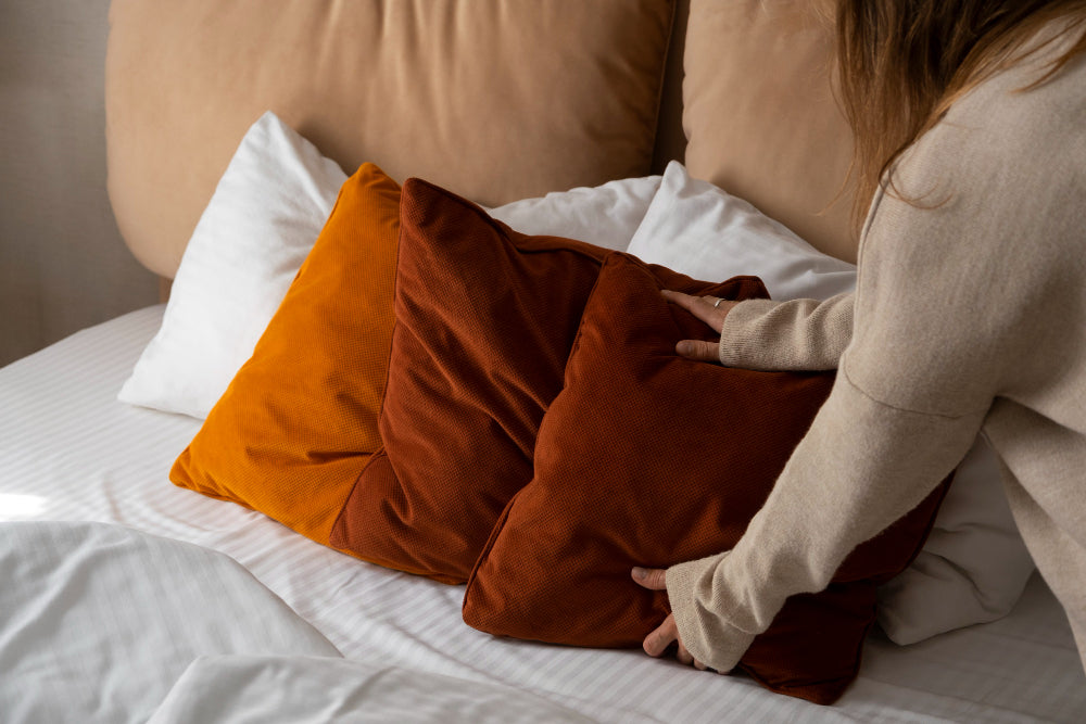 A person arranging burnt orange throw pillows on a neatly made bed with white sheets, showing how to layer bedding with accent cushions for a cozy, stylish look.