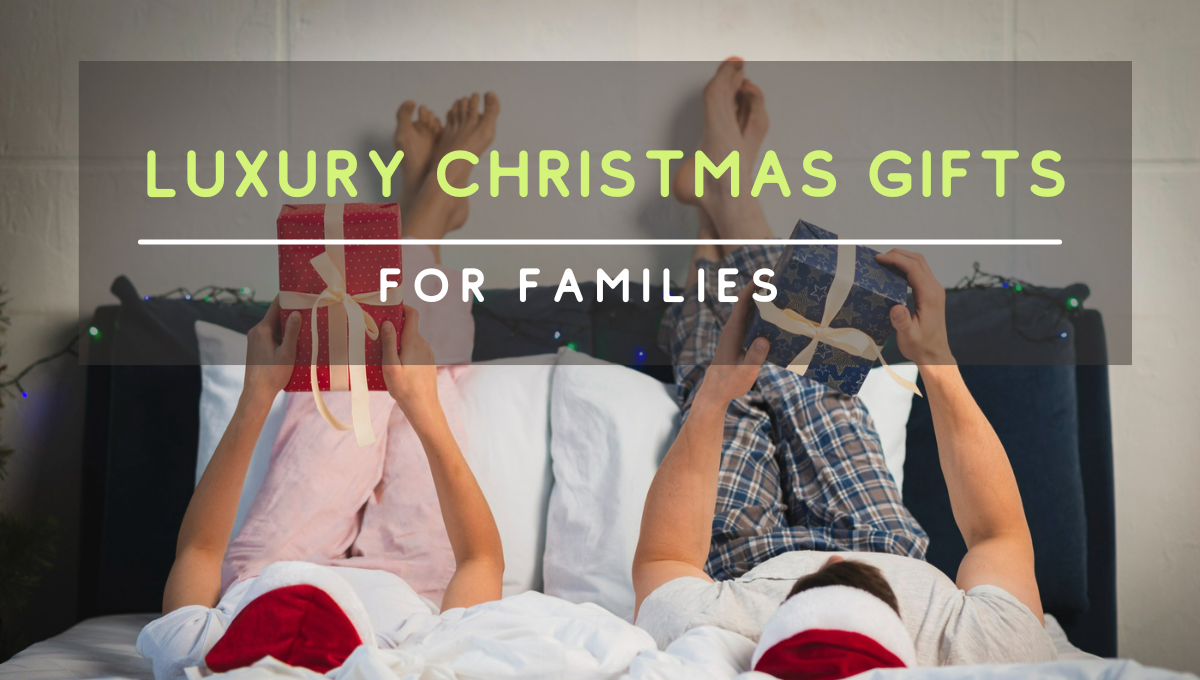 Two people wearing Christmas hats lie on a bed with their feet up, each holding a wrapped holiday gift, with the text “Luxury Christmas Gifts for Families” displayed across the image.