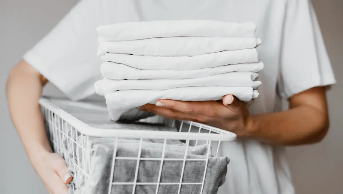 Person holding a stack of freshly washed white bed sheets above a laundry basket.