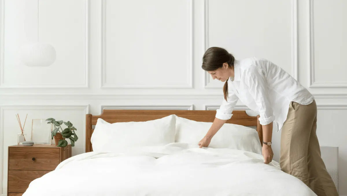 A woman smoothing white bedding in a calm, minimal bedroom while making the bed.