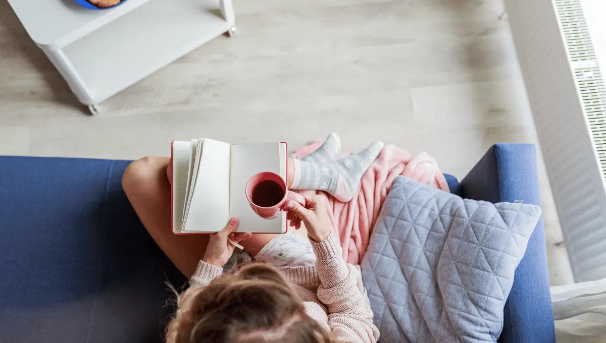 Person relaxing at home with a book, tea, and cozy blanket, representing simple daily habits for wellness and comfort.