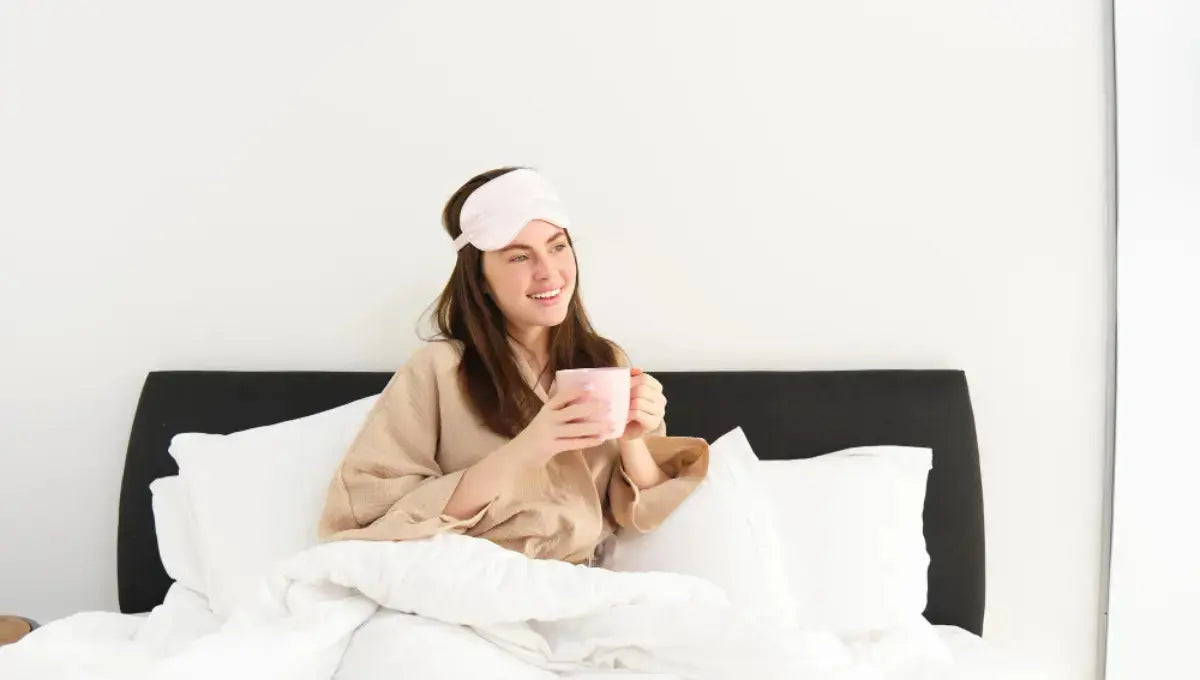 Woman sitting in bed with a sleep mask holding a warm drink during a relaxed morning routine.