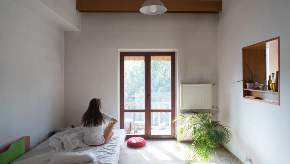 Person sitting on an unmade bed in a bright bedroom with open windows during hot weather without air conditioning.