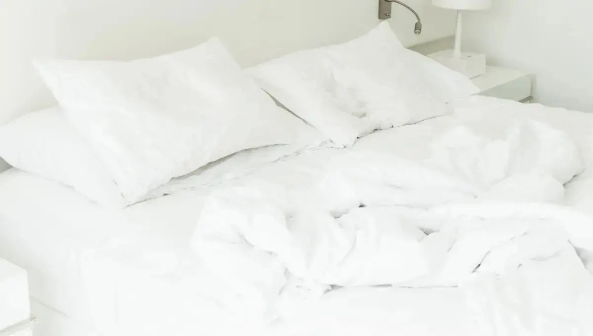 White comforter and pillows on an unmade bed in a bright bedroom.