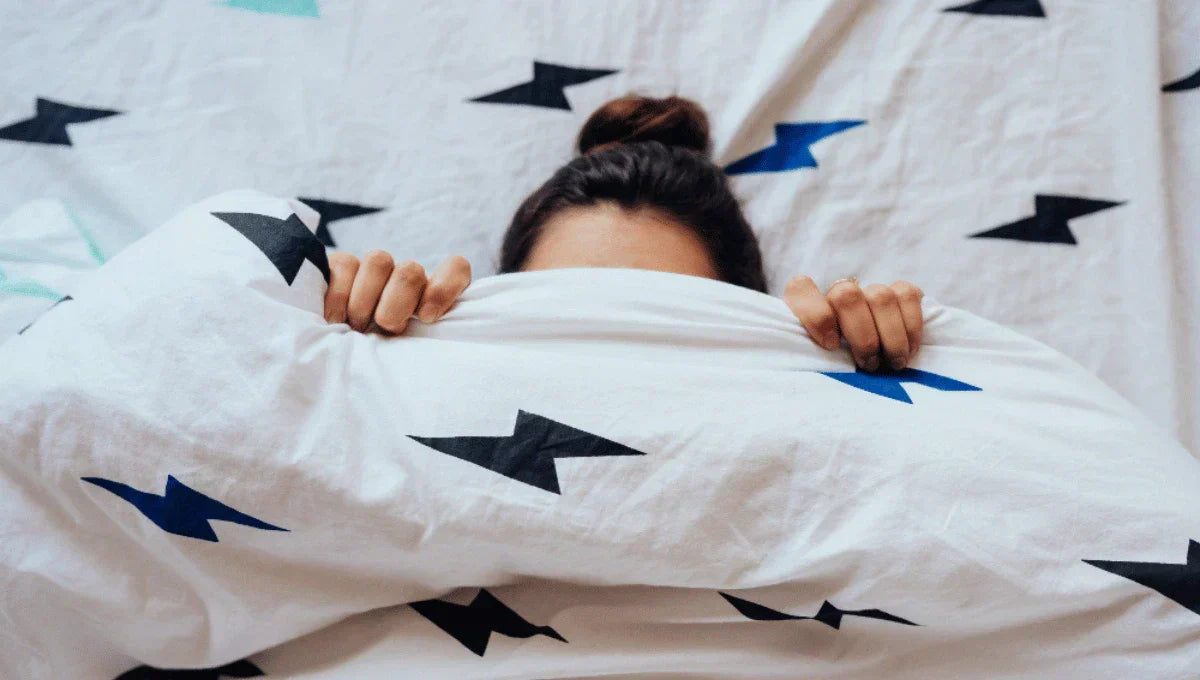 Woman peeking over lightning-patterned duvet in bed, symbolizing sleep awareness and restless nights for World Sleep Day.