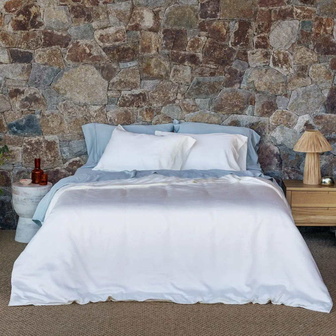 A neatly made bed with white bedding and light blue pillows and duvet, set against a rustic stone wall with a wooden nightstand and lamp on each side. 