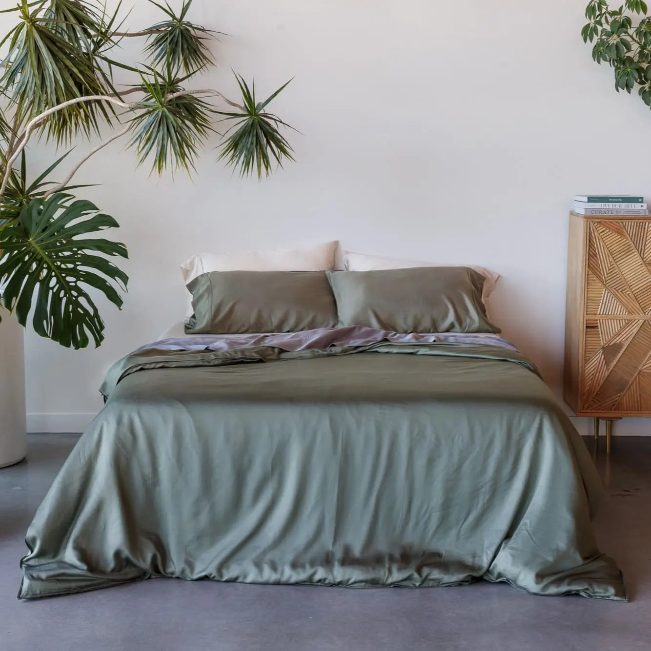 Bed with green bedding in a room with plants and a wooden side table. 